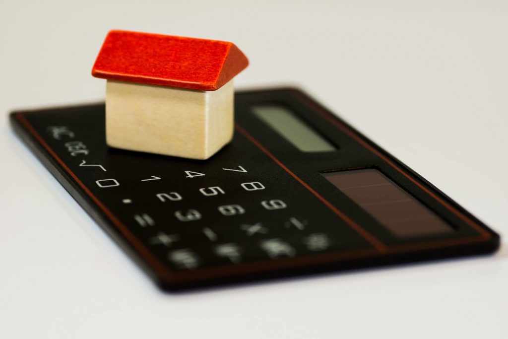 Ein Bild mit einem kleinen Holzspielzeughaus auf einem Taschenrechner, das den finanziellen Aspekt von Wohneigentum und die Verbindung zwischen Berechnungen und dem Konzept von Zuhause symbolisiert.